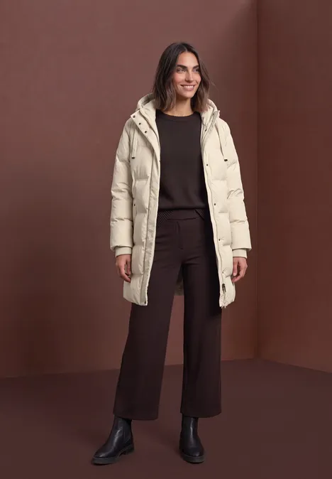 Frau in beigefarbener Steppjacke, braunem Outfit und schwarzen Stiefeletten vor braunem Hintergrund.