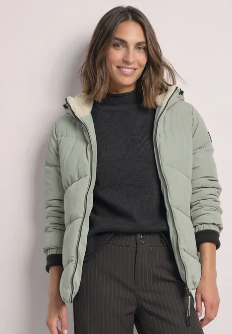 Puffer Jacke mit Teddyfutter in der Kapuze Fresh Sage Frau mit grüner Steppjacke und schwarzem Pullover lächelt vor hellem Hintergrund.