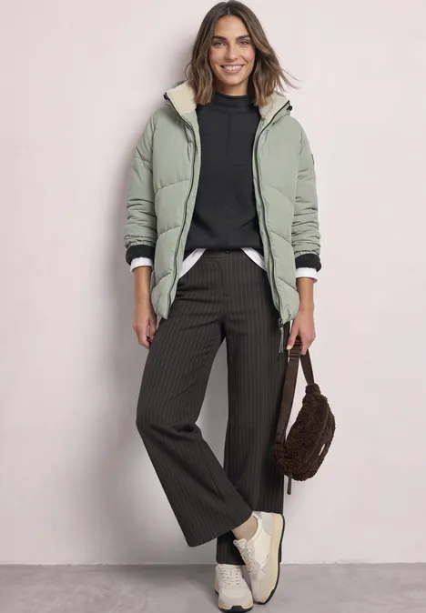 Puffer Jacke mit Teddyfutter in der Kapuze Fresh Sage Frau in grüner Jacke mit Teddyfutter, schwarzem Pullover, weiter Hose und weißen Sneakern, braune Tasche.