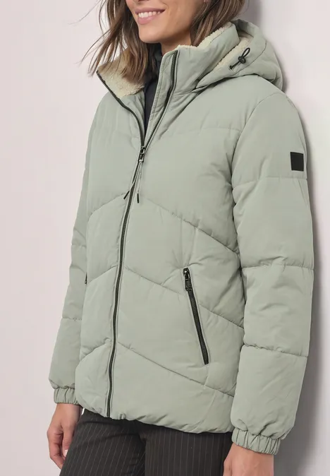 Puffer Jacke mit Teddyfutter in der Kapuze Fresh Sage Eine Frau trägt eine hellgrüne, wattierte Jacke mit Kapuze und Reißverschlusstaschen.