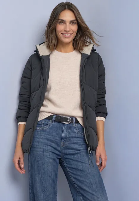 Frau in schwarzer Steppjacke mit Kapuze, beigem Pullover und blauen Jeans, lächelnd.