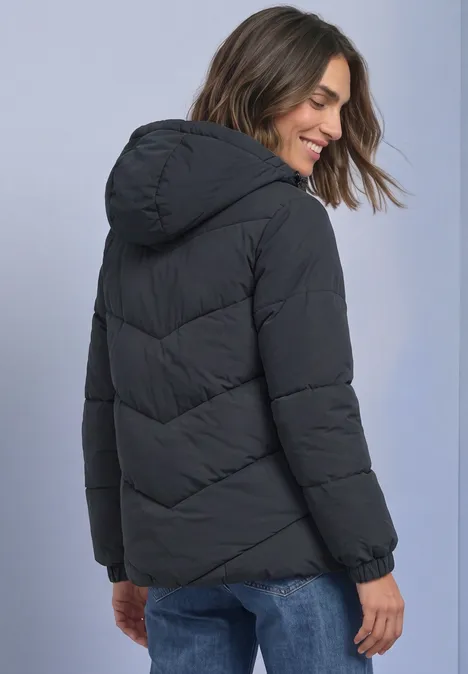 Frau in schwarzer Steppjacke mit Kapuze, blickt über die Schulter und lächelt.