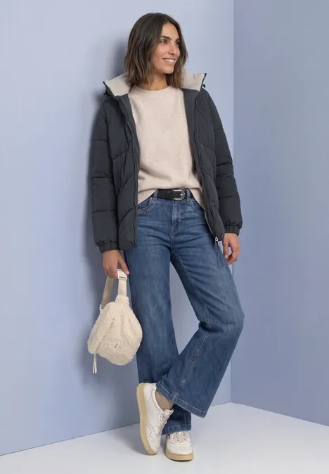 Frau in Steppjacke, Pullover, weiter Jeans, Plateauschuhen und Stricktasche vor lila Wand