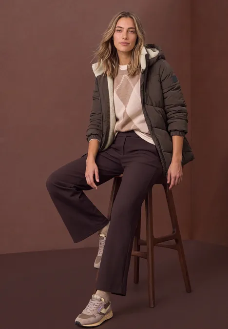 Puffer Jacke mit Teddyfutter in der Kapuze auburn brown Frau sitzt auf Hocker vor braunem Hintergrund, trägt braune Steppjacke, Pullover, weite dunkle Hose und Sneaker.
