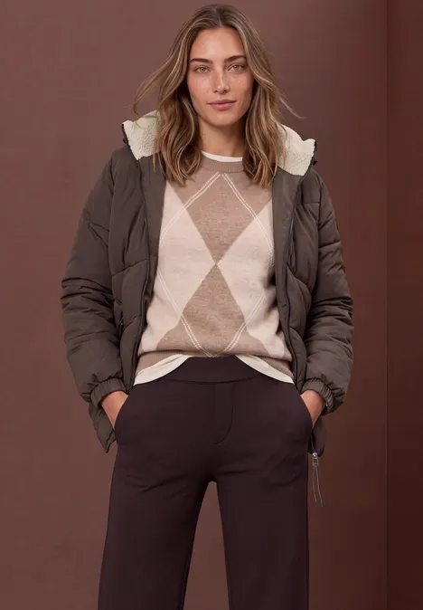 Puffer Jacke mit Teddyfutter in der Kapuze auburn brown Frau mit brauner Steppjacke und beigem Argyle-Pullover, dunkle Hose, Hände in den Taschen.