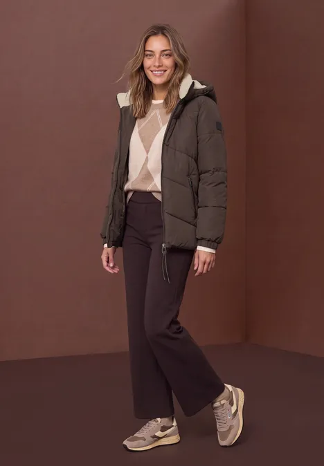 Puffer Jacke mit Teddyfutter in der Kapuze auburn brown Frau im braunen Steppmantel, Pullover, dunkler Hose und Sneakern vor braunem Hintergrund, lächelnd.