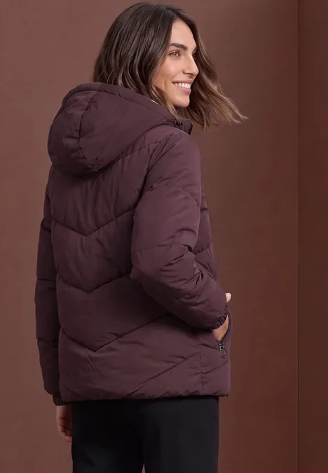 Eine Frau trägt eine burgunderfarbene Steppjacke mit Kapuze und lächelt seitlich zur Kamera.