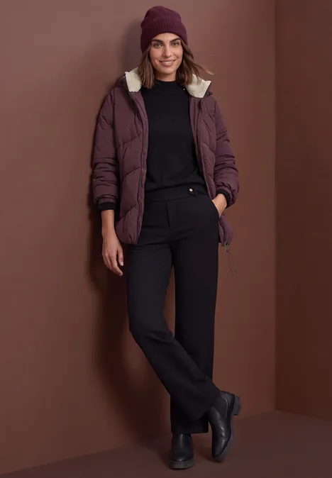 Frau in beerenfarbener Jacke, schwarzem Rollkragenpullover, schwarzer Hose, Boots & Mütze vor braunem Hintergrund.