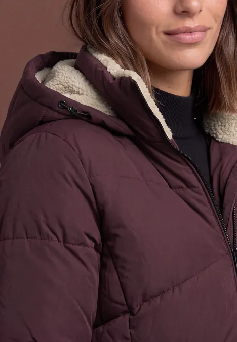 Eine Frau trägt eine wattierte, weinrote Kapuzenjacke mit hellem Teddyfellfutter.