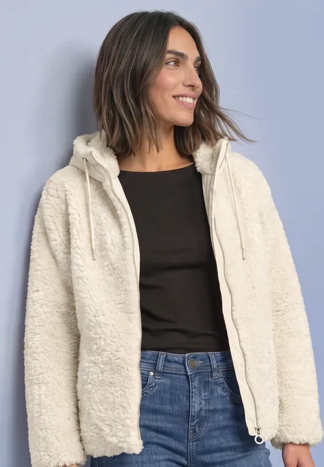 Frau in weißer Teddy-Fleecejacke mit Kapuze, schwarzem Shirt und Jeans, lächelnd zur Seite blickend.