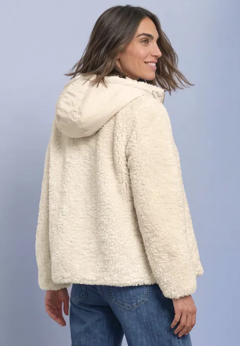 Frau in cremefarbener Teddyjacke mit Kapuze und Jeans, leicht seitlich von hinten.