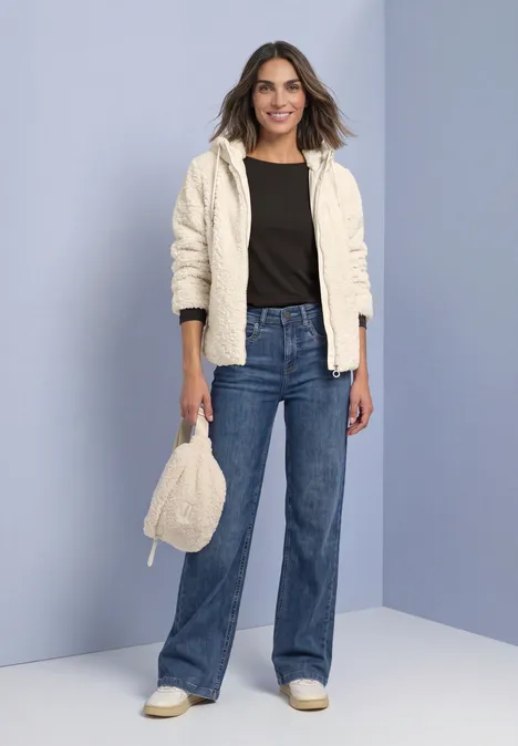 Frau in cremefarbener Kapuzenfleecejacke, schwarzem Top, weiter blauer Jeans, weißen Sneakern und passender Tasche.