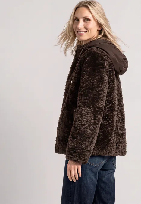 Eine blonde Frau trägt seitlich stehend eine flauschige, braune Kapuzenjacke und lächelt.