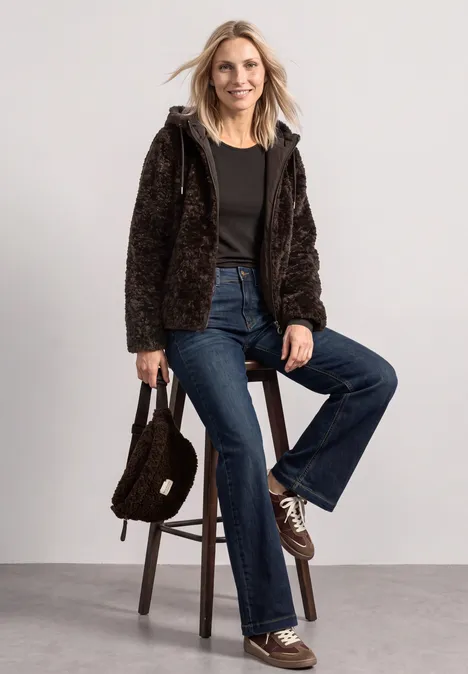 Frau in brauner Teddyfelljacke, schwarzem Shirt und Schlagjeans sitzt auf Hocker, hält passende Handtasche.