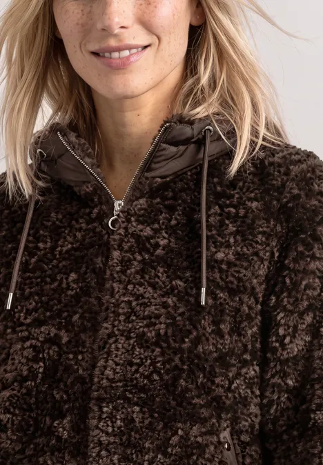 Frau mit Sommersprossen trägt dunkelbraune, flauschige Teddyjacke mit Kapuze und Reißverschluss und lächelt dezent.