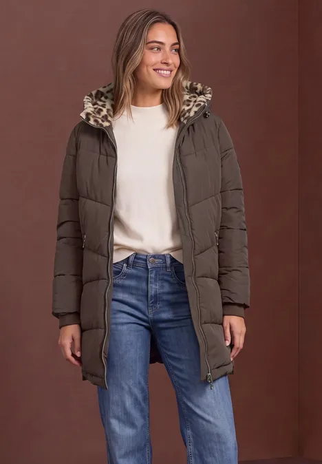 Frau in braunem Steppmantel mit Kunstfellkapuze, weißem Pullover und Jeans, lächelnd vor braunem Hintergrund.
