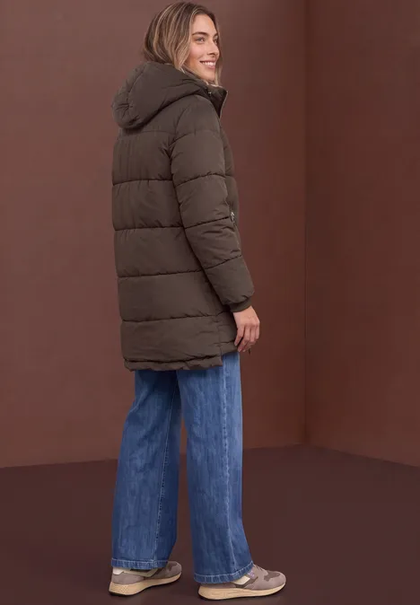 Frau mit brauner Steppjacke und Kapuze, blauen Jeans und Sneakern steht seitlich vor braunem Hintergrund.
