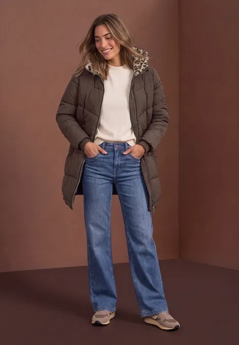 Frau in brauner Steppjacke, weißem Pullover und blauen Jeans, lächelnd vor braunem Hintergrund.