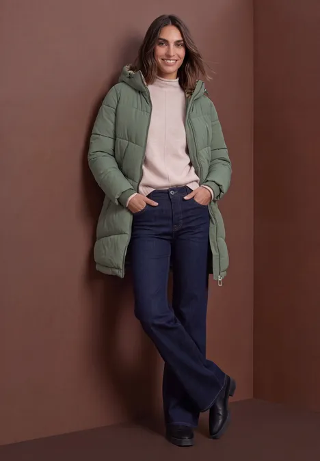 Gevoerde parka met nepbont in luipaardprint Somber Green Frau vor braunem Hintergrund, in grünem Steppmantel mit Kapuze, rosa Pullover, Jeans und schwarzen Schuhen, lächelt.