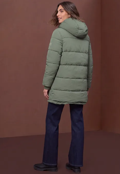 Gevoerde parka met nepbont in luipaardprint Somber Green Frau in grüner Steppjacke, blauen Jeans und schwarzen Schuhen, steht vor braunem Hintergrund und blickt zurück.