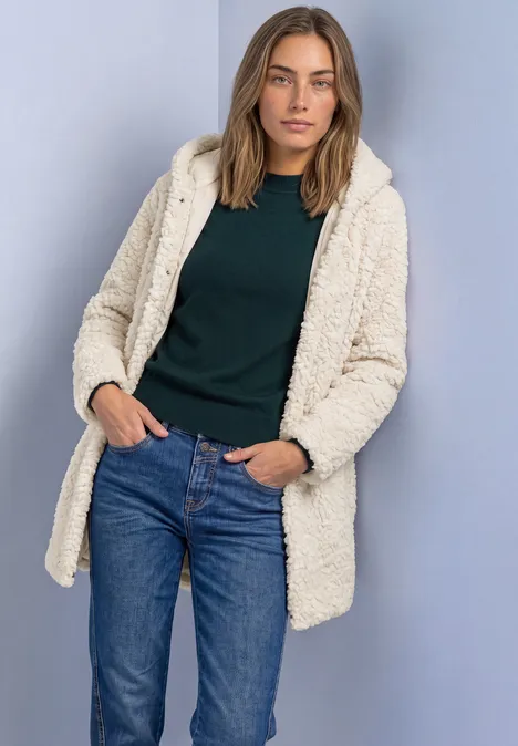 Une femme porte un manteau teddy crème, un haut vert foncé et un jean bleu sur fond neutre.