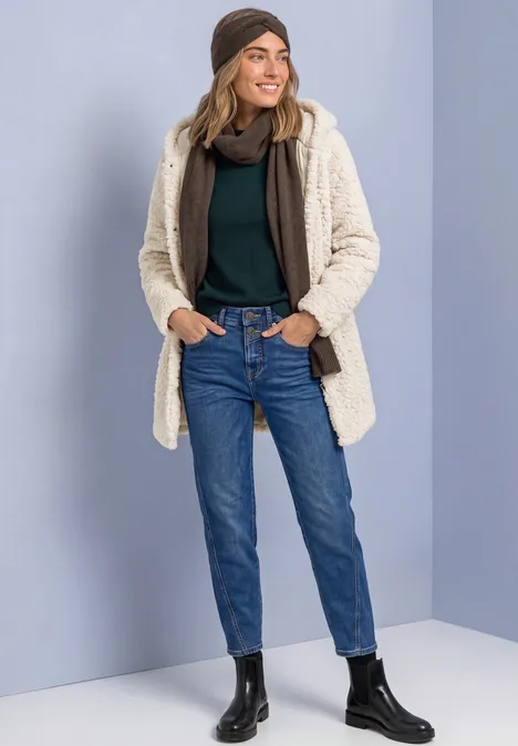 Femme en jean, pull à col roulé noir, bonnet, veste molletonnée crème et bottines noires souriante.
