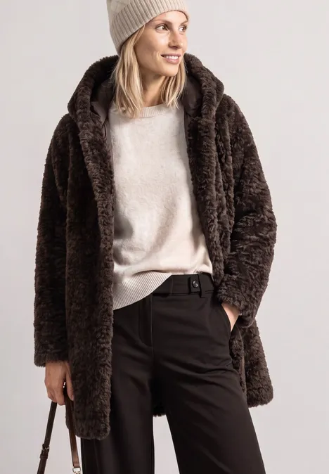 Femme en manteau teddy marron, pull crème, pantalon noir et bonnet, tenant une petite sacoche.