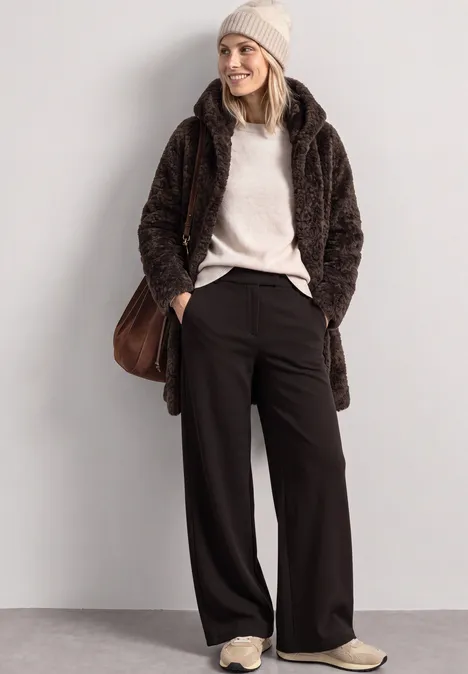 Femme en manteau teddy marron, pull beige, pantalon large noir, bonnet crème et baskets.
