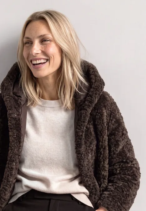 Une femme blonde souriante porte une veste marron en moumoute et un t-shirt clair sur fond neutre.
