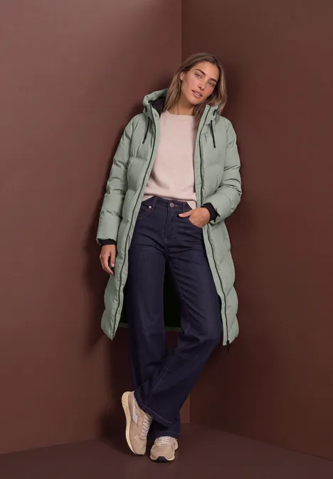 Veste matelassée à capuche et détails zippés Fresh Sage Femme en doudoune longue vert menthe, jean ample et baskets, posant devant un mur marron.