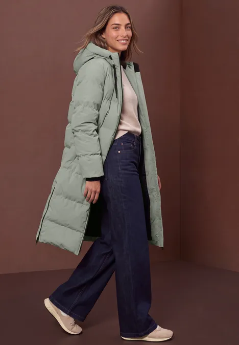 Veste matelassée à capuche et détails zippés Fresh Sage Femme portant un long manteau doudoune vert menthe, chemisier clair et jean large, sur fond marron.