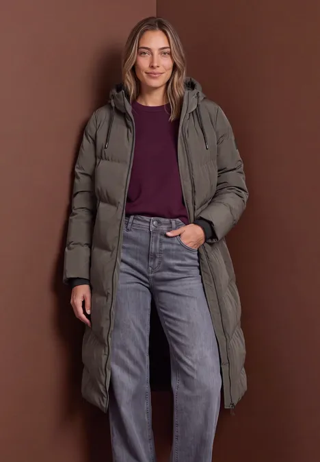 Femme portant un manteau matelassé long, un pull et un jean devant un fond marron.
