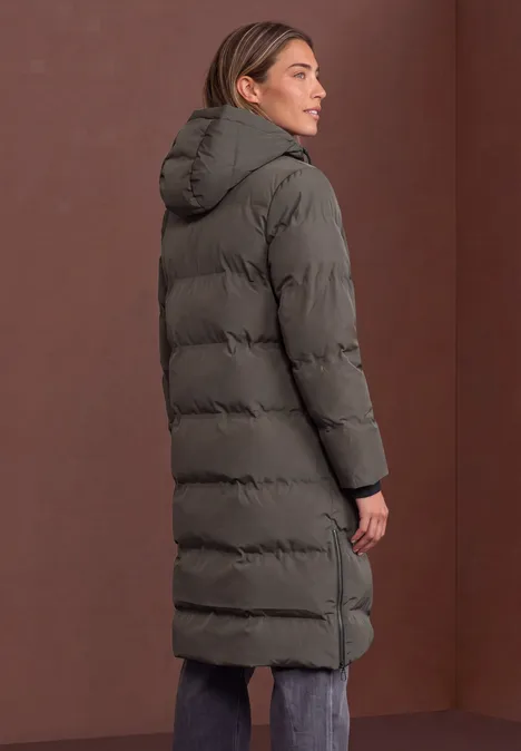 Femme de profil portant un long manteau matelassé vert foncé avec capuche devant un fond brun.