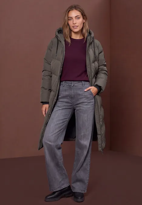 Femme portant un long manteau matelassé, un haut bordeaux et un jean gris large sur fond marron.