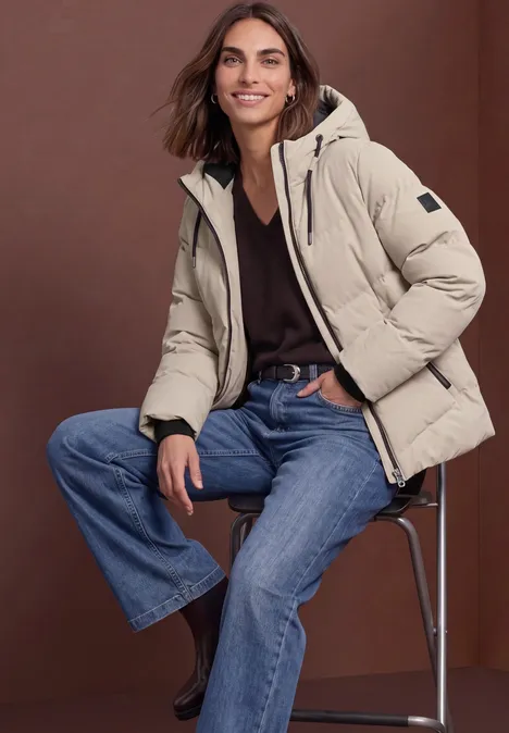 Pufferjacke mit Kapuze barley beige Frau trägt beige Steppjacke, schwarzen Pullover und Jeans, sitzt auf Hocker.