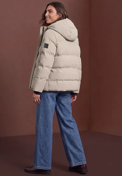 Pufferjacke mit Kapuze barley beige Frau in beige Daunenjacke mit Kapuze und blauen Jeans, seitlich stehend, blickt über die Schulter