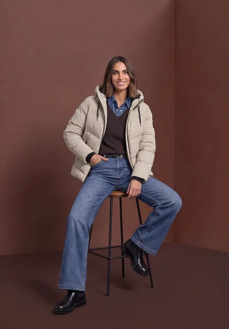 Pufferjacke mit Kapuze barley beige Frau in beiger Steppjacke, Hemd und Jeans sitzt lässig auf einem Hocker vor braunem Hintergrund.