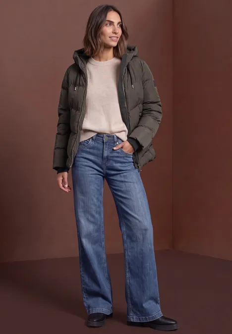 Vrouw in donkergroene gewatteerde jas, beige trui en wijde blauwe jeans voor een bruine achtergrond.