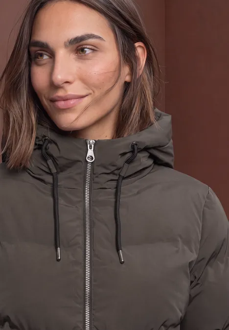 Close-up van een vrouw in een bruine gewatteerde jas met capuchon, ritssluiting en koordjes.