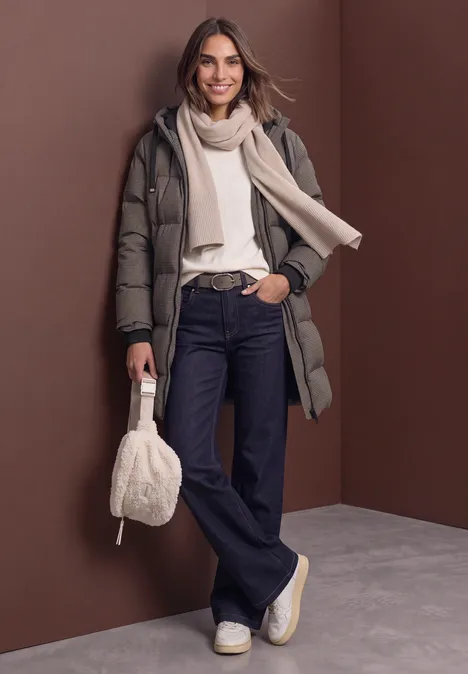 Frau in langer Daunenjacke, beigem Pullover, Schal, Jeans, Sneakern, hält flauschige Tasche vor braunem Hintergrund.