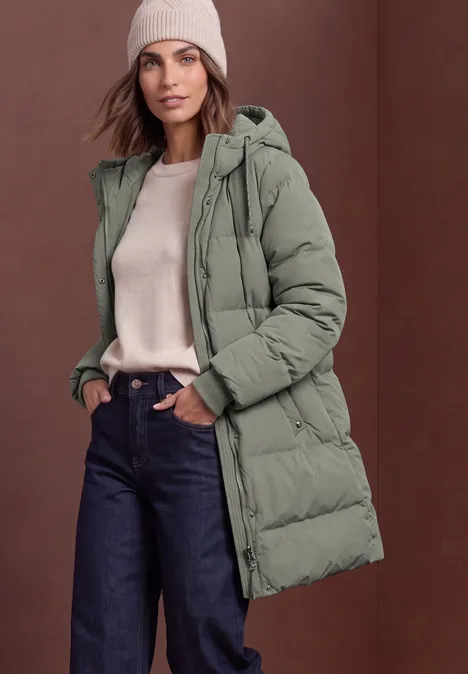Lange Jacke mit Kapuze und Steppmuster Somber Green Eine Frau trägt einen grünen Steppmantel mit Kapuze, beigen Pullover, Jeans und Mütze vor neutralem Hintergrund.