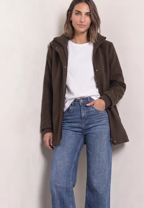 Frau in weißem Shirt, blauer Jeans und braunem Mantel vor hellem Hintergrund