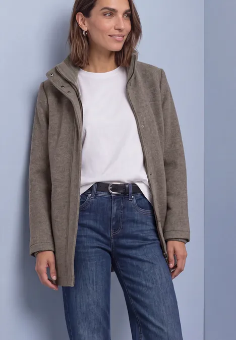 Frau in weißem T-Shirt, blauen Jeans und grauer Kapuzenjacke steht vor einfarbigem Hintergrund.