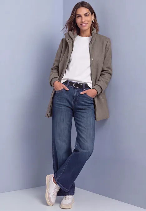 Frau in grauem Mantel, weißem Shirt, blauen Jeans und weißen Sneakern steht entspannt, lehnt an Wand.