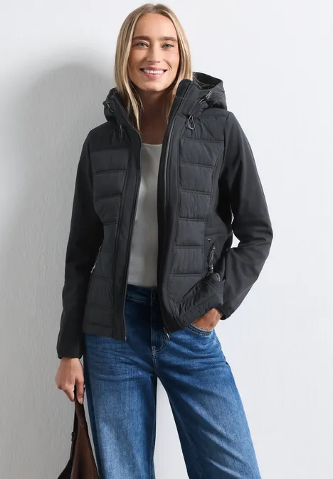 Lächelnde Frau in schwarzer Kapuzen-Steppjacke, blauer Jeans und Tasche vor weißem Hintergrund.