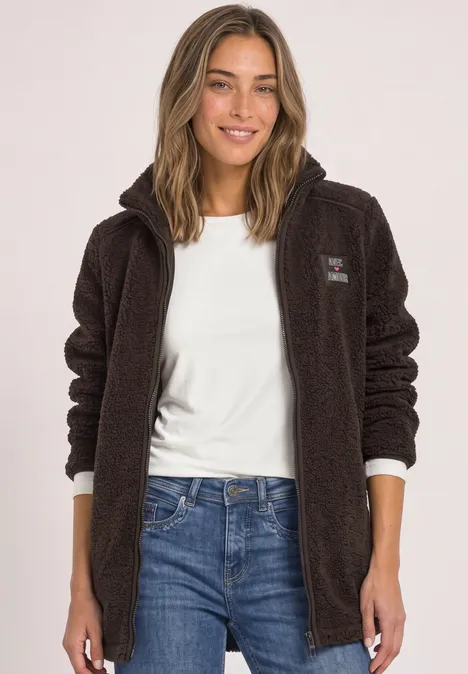 Frau trägt braune Teddy-Fleecejacke, weiße Bluse und Jeans, lächelnd.