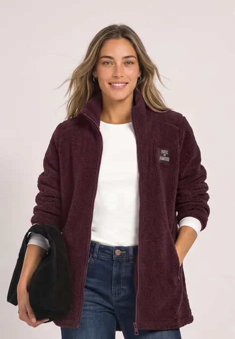 Frau trägt weinrote Fleecejacke, weiße Bluse, Jeans und hält schwarze Tasche.