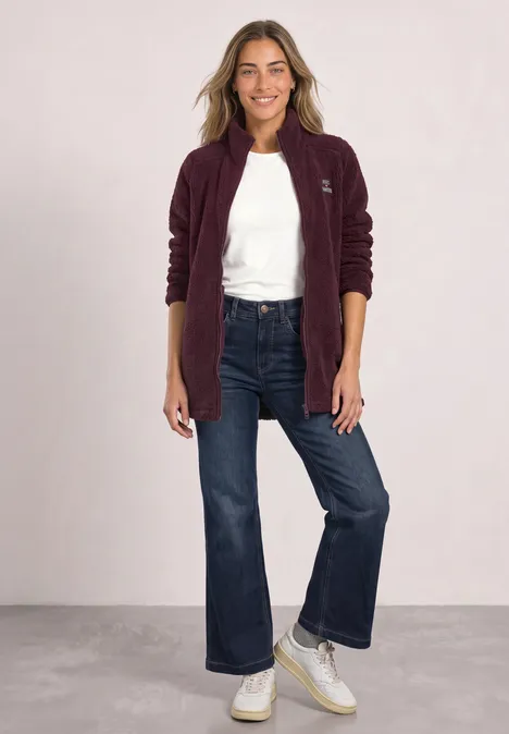 Eine lächelnde Frau in weinroter Fleecejacke, weißem T-Shirt, weiten Jeans und weißen Sneakers vor neutralem Hintergrund.
