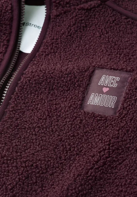 Nahaufnahme einer weinroten Fleecejacke mit Logo-Patch 'AVEC AMOUR' und Reißverschluss.