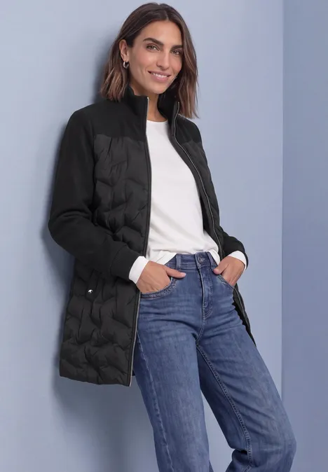 Frau in schwarzer Steppjacke, weißem Oberteil und blauen Jeans, lächelnd mit Händen in den Taschen vor grauem Hintergrund.